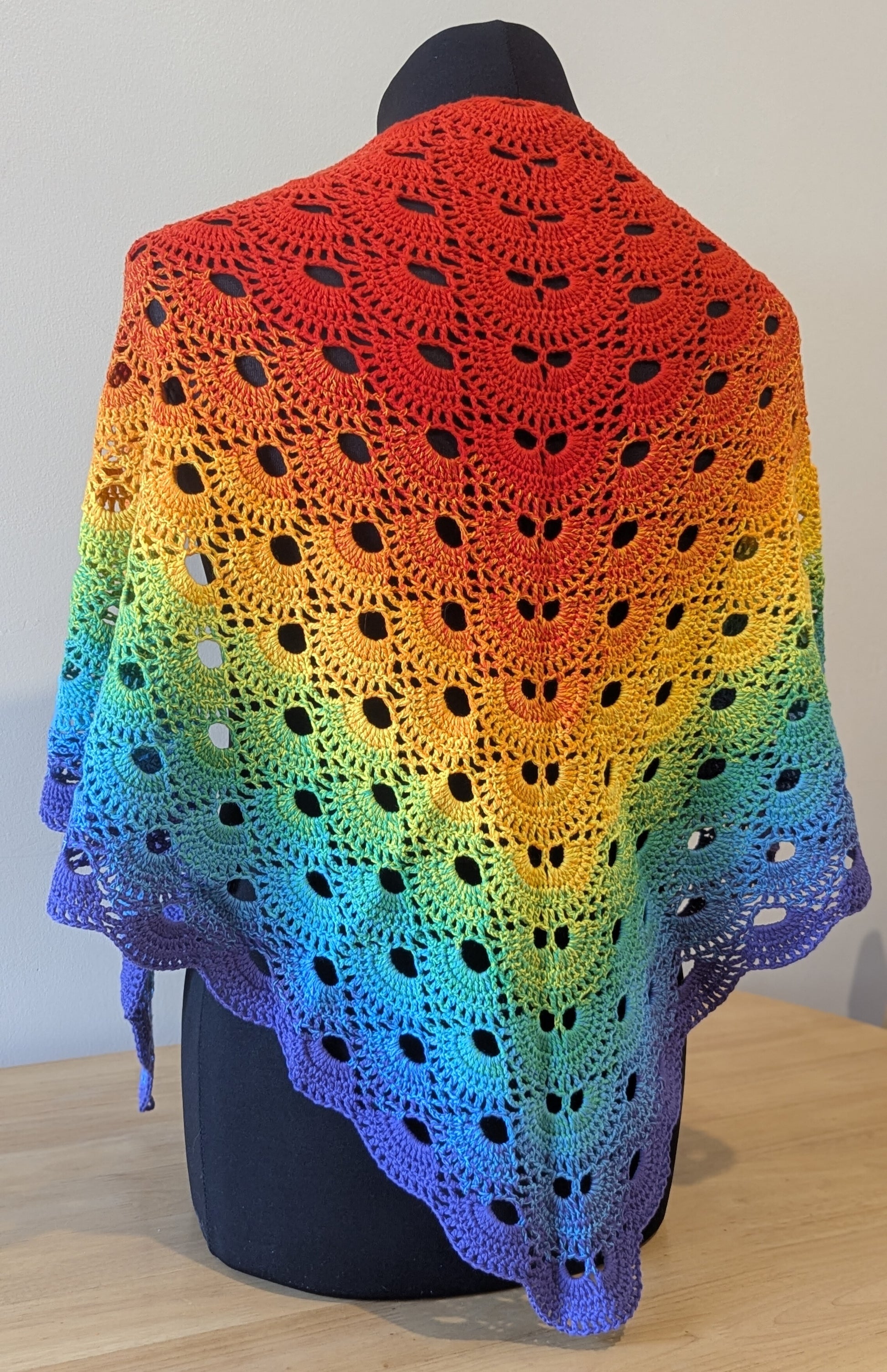 Colourful crocheted shawl with a rainbow gradient on a mannequin against a plain background.