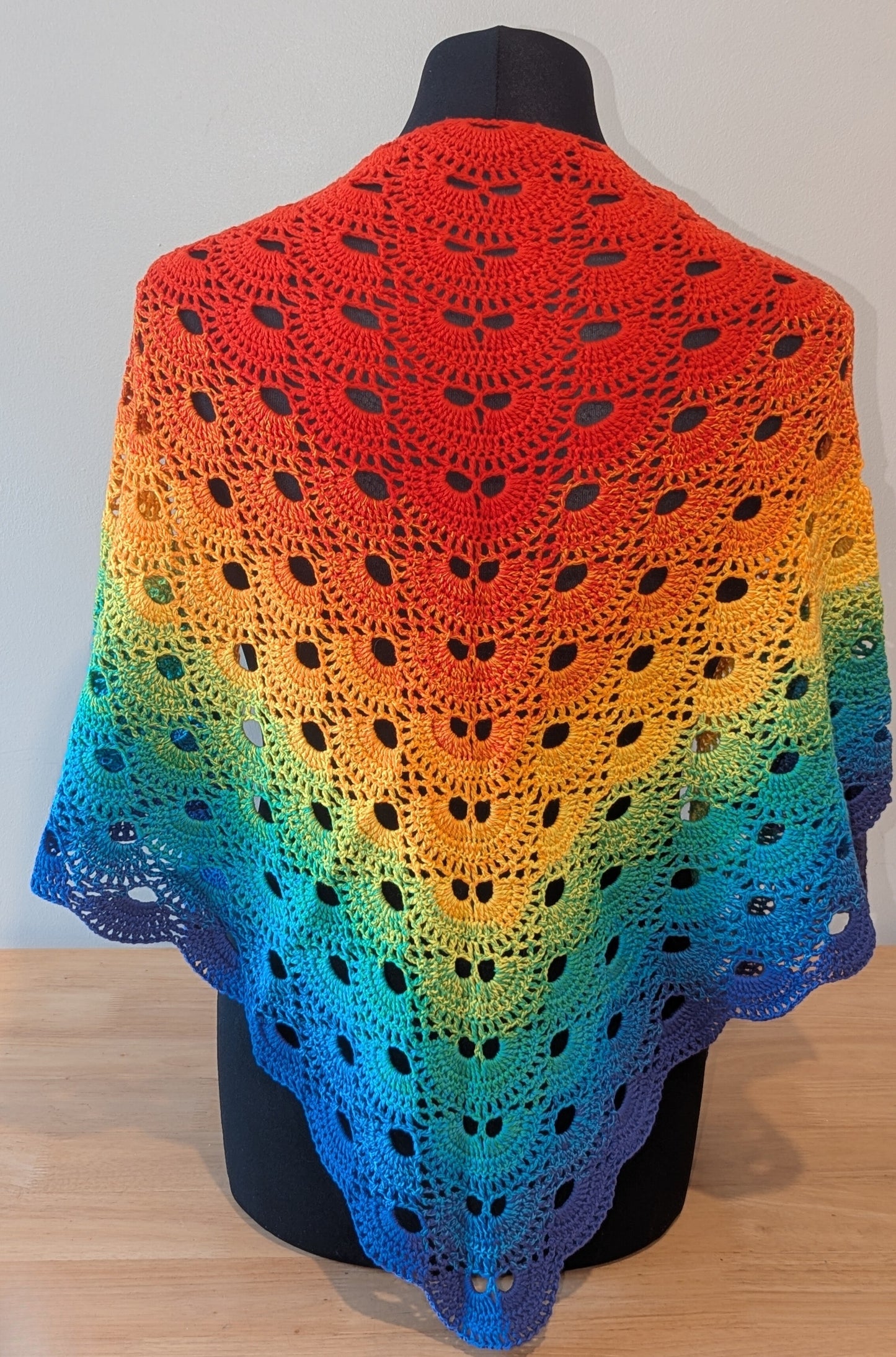 Colorful crocheted shawl with a gradient pattern on a mannequin against a plain background.