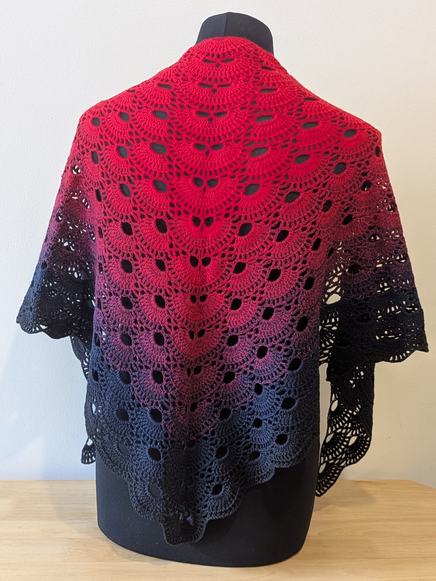 Crocheted shawl with a gradient of red to black on a mannequin against a plain background