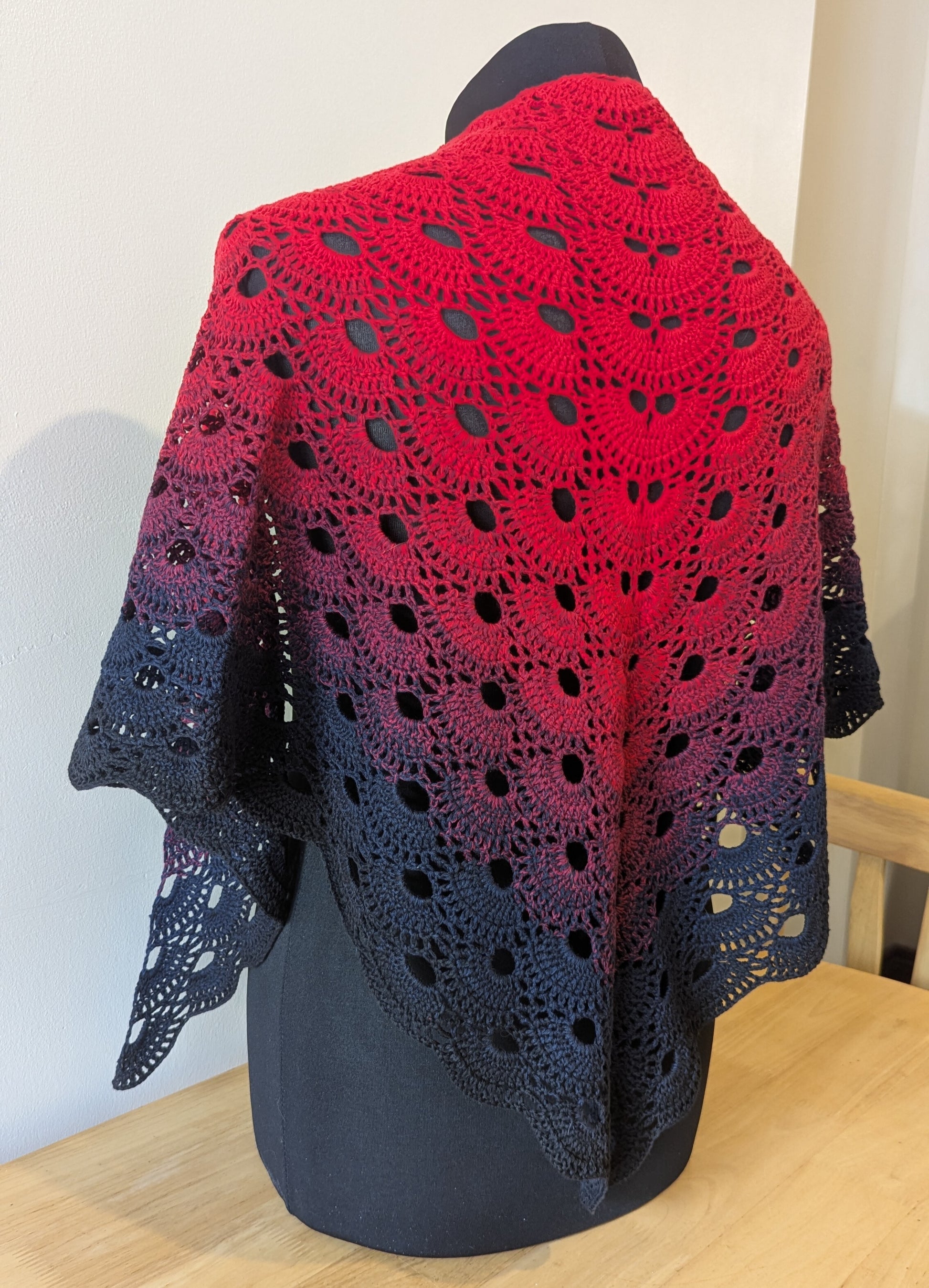 Crocheted shawl with a gradient from red to black on a mannequin against a white wall.