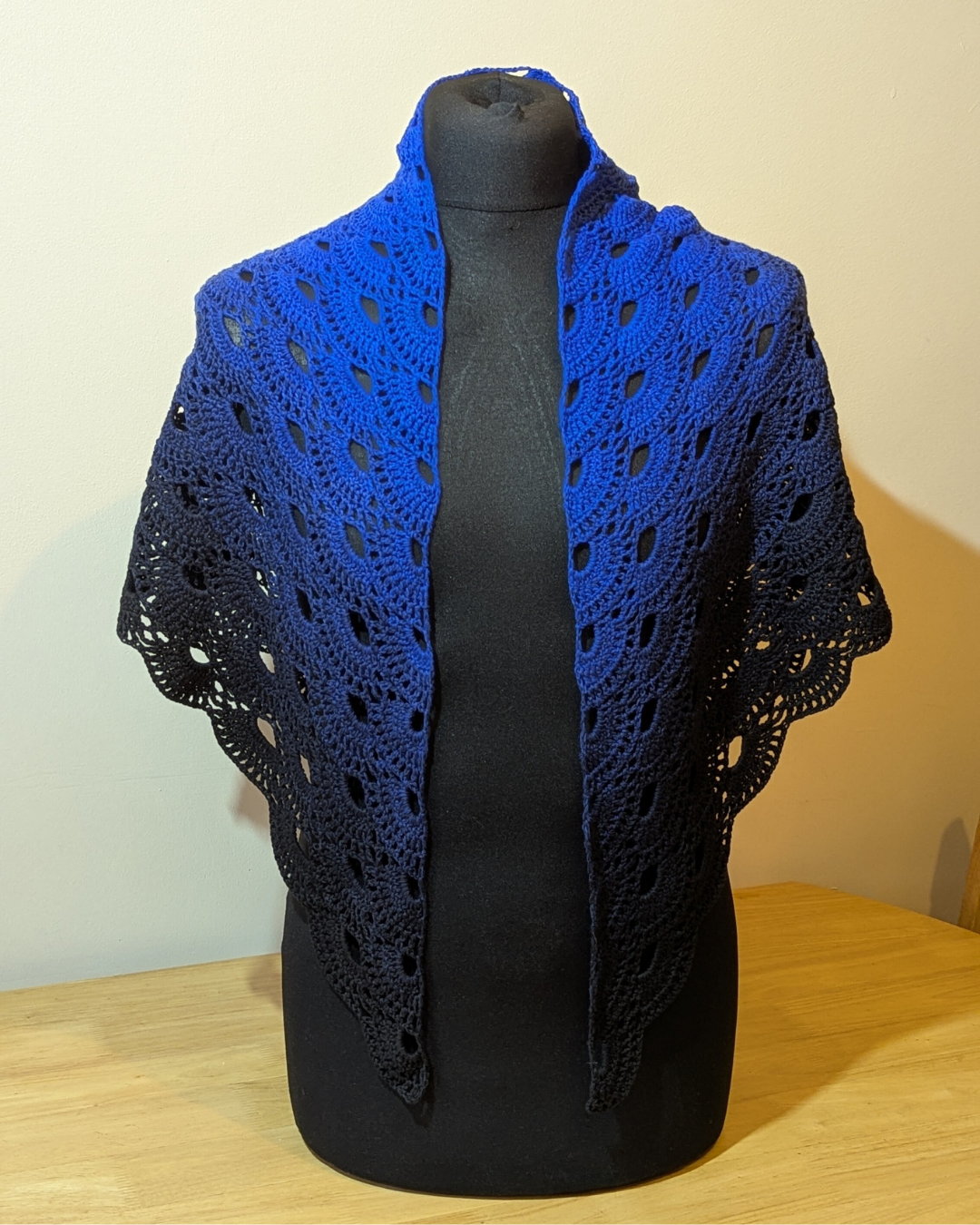 Blue crocheted shawl on a black mannequin against a beige wall.