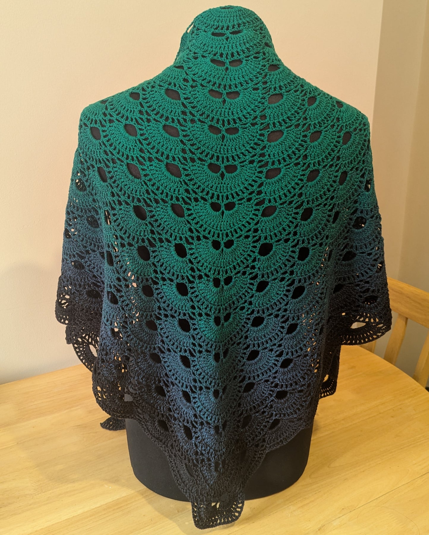 Crocheted shawl with a gradient from green to black draped over a mannequin on a wooden table.