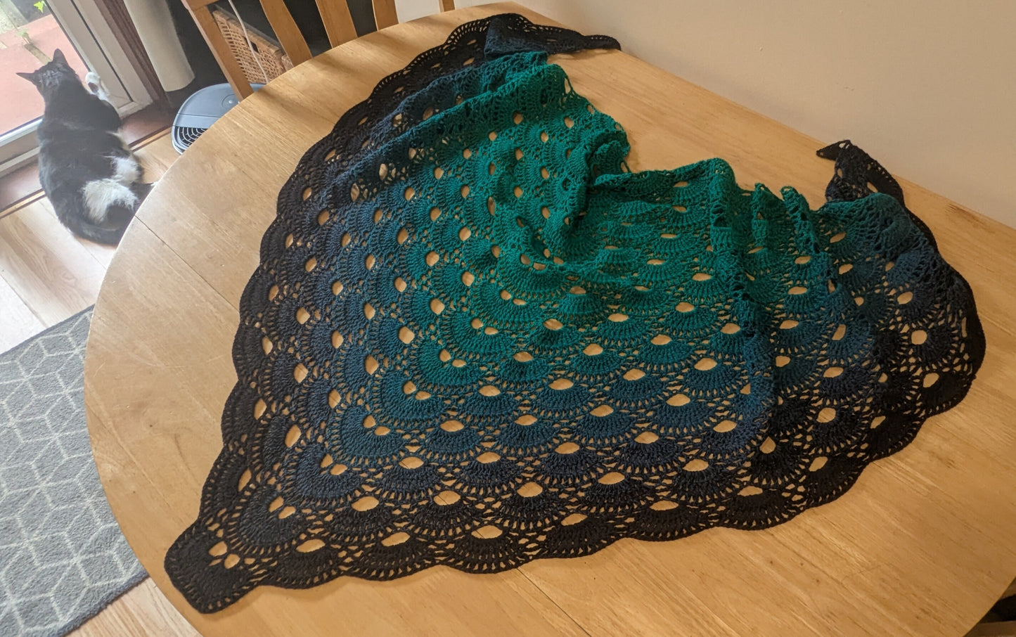 Green and black lacey shawl on a wooden table with a cat in the background.